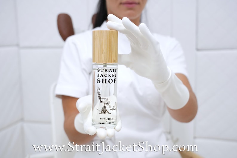 May include: A clear glass spray bottle with a light wood cap. The label features the black text STRAIT JACKET SHOP, a graphic illustration of a vintage dental chair, and the text MEMORIES Dental Office printed in black on the front surface.
