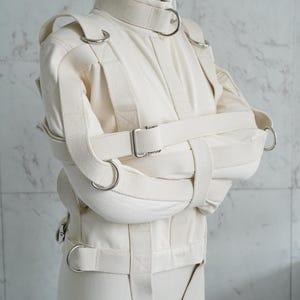 May include: A white canvas straitjacket with multiple straps and buckles. The jacket is designed to restrain the arms and torso. The jacket has metal rings and a buckle closure.