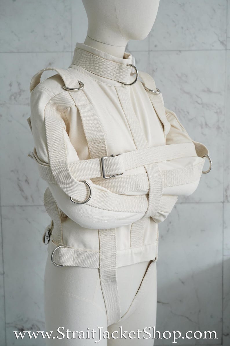 May include: A white canvas straitjacket with multiple straps and buckles. The jacket is designed to restrain the arms and torso. The jacket has metal rings and a buckle closure.