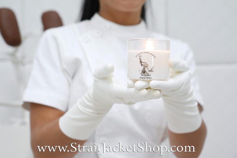 May include: A clear glass candle container with white wax and a lit wick. The label features a black line drawing of a dental chair with the text STRAITJACKETSHOP, MEMORIES, and Dental Office printed in black ink on the front of the glass.