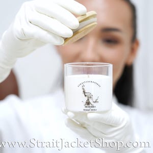 May include: A clear glass candle jar filled with white wax, featuring a black graphic of a dental chair and the text STRAITJACKETSHOP MEMORIES Dental Office. The jar includes a round, light-colored wooden lid held above the opening.