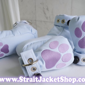 May include: Light blue padded mitts featuring purple paw print appliques on the palms. Each mitt includes a wrist strap with silver-toned metal eyelets and a D-ring closure. The text www.StraitJacketShop.com is printed at the bottom of the image.