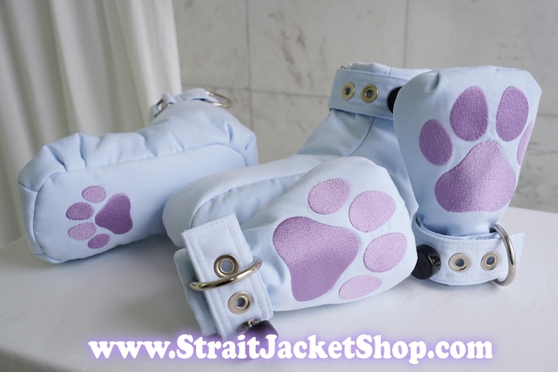 May include: Light blue padded mitts featuring purple paw print appliques on the palms. Each mitt includes a wrist strap with silver-toned metal eyelets and a D-ring closure. The text www.StraitJacketShop.com is printed at the bottom of the image.