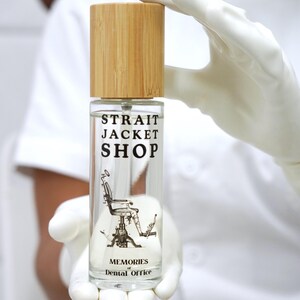 May include: A clear glass spray bottle with a light brown bamboo cap. The label features the black text STRAIT JACKET SHOP, a graphic illustration of a vintage dental chair, and the text MEMORIES of Dental Office. The bottle is cylindrical.