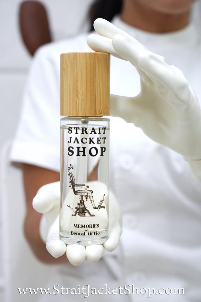 May include: A clear glass spray bottle with a light brown bamboo cap. The label features the black text STRAIT JACKET SHOP, a graphic illustration of a vintage dental chair, and the text MEMORIES of Dental Office. The bottle is cylindrical.