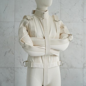 May include: A white canvas straitjacket with straps and metal rings. The jacket is worn by a white mannequin. The text "www.StraitJacketShop.com" is visible at the bottom of the image.