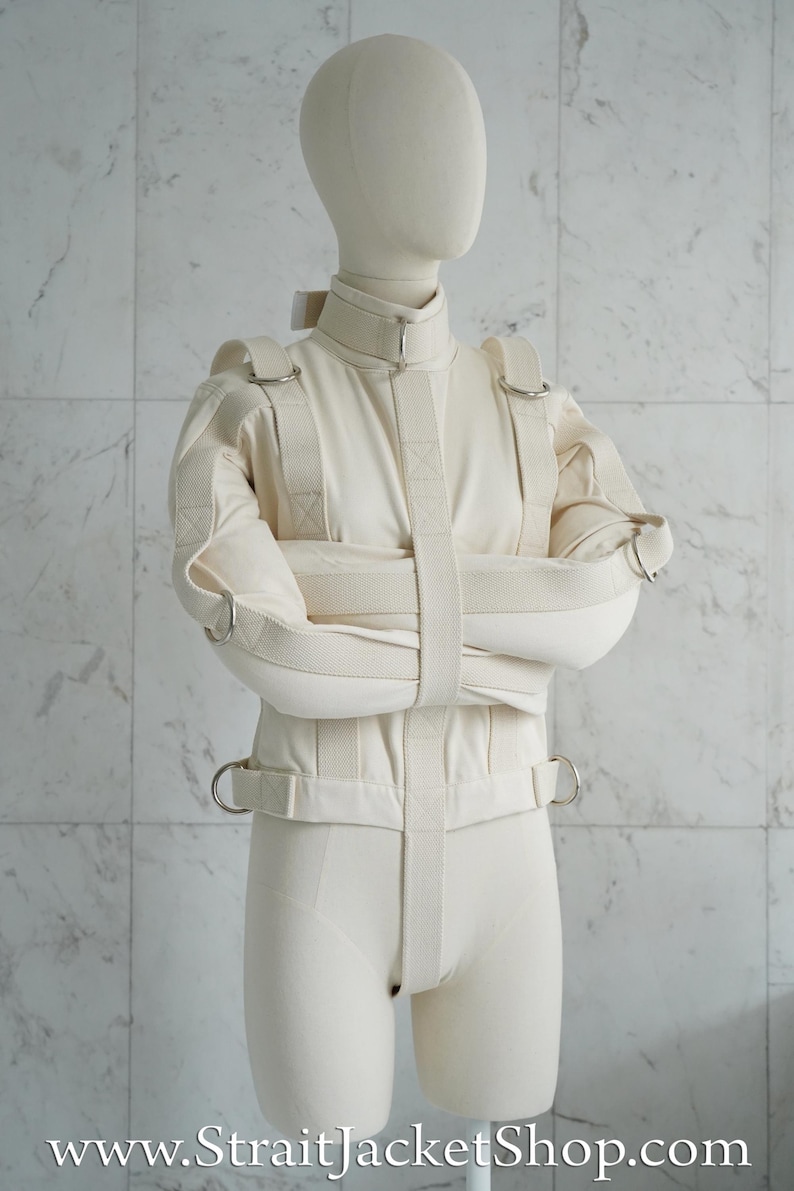 May include: A white canvas straitjacket with straps and metal rings. The jacket is worn by a white mannequin. The text "www.StraitJacketShop.com" is visible at the bottom of the image.