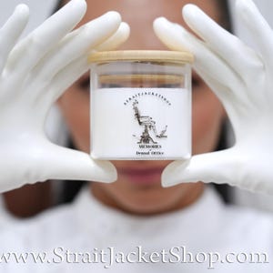 May include: A clear glass candle jar with a light wood lid, filled with white wax. The label features a black line drawing of a dental chair with the text STRAITJACKETSHOP, MEMORIES, and Dental Office printed in black ink on the front surface.