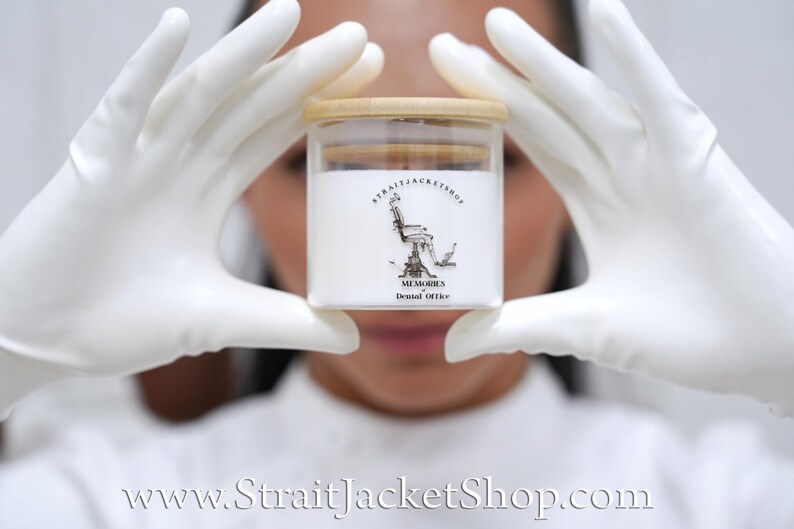 May include: A clear glass candle jar with a light wood lid, filled with white wax. The label features a black line drawing of a dental chair with the text STRAITJACKETSHOP, MEMORIES, and Dental Office printed in black ink on the front surface.