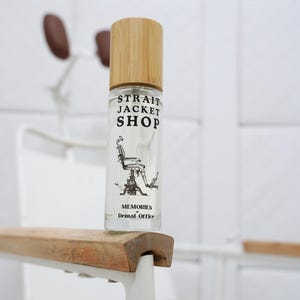 May include: A clear glass roller bottle with a light wood cap. The label features the black text STRAIT JACKET SHOP, an illustration of a vintage dental chair, and the text MEMORIES of Dental Office. The bottle is positioned on a wooden armrest.