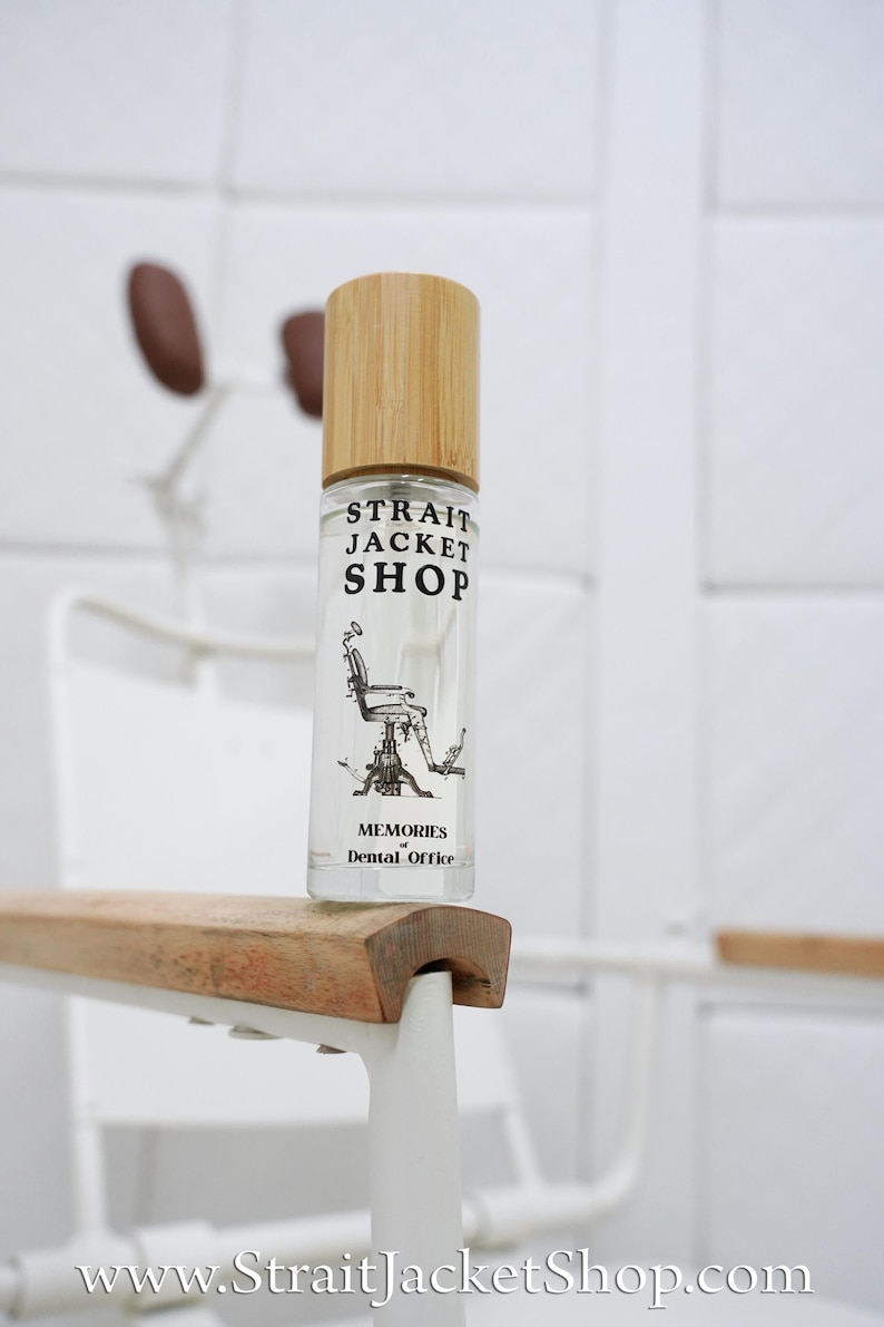 May include: A clear glass roller bottle with a light wood cap. The label features the black text STRAIT JACKET SHOP, an illustration of a vintage dental chair, and the text MEMORIES of Dental Office. The bottle is positioned on a wooden armrest.