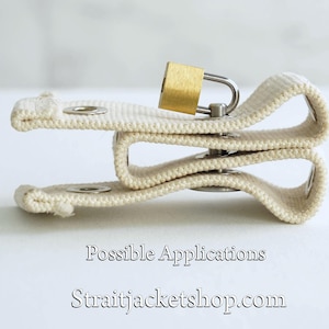 May include: A white canvas strap with a metal buckle and a small gold padlock. The strap is folded in half and the padlock is attached to the strap. The text "Possible Applications" and "straitjacketshop.com" is printed below the strap.