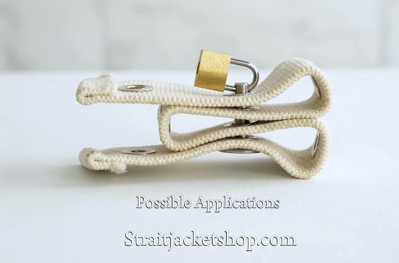 May include: A white canvas strap with a metal buckle and a small gold padlock. The strap is folded in half and the padlock is attached to the strap. The text "Possible Applications" and "straitjacketshop.com" is printed below the strap.