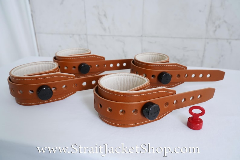 May include: Four brown leather wrist restraints with white lining and black metal buckles. The restraints are laid out on a white surface. A red plastic key is also visible in the image.