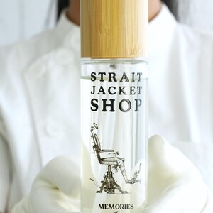 May include: A clear glass spray bottle with a light-coloured bamboo cap. The label features the text "STRAIT JACKET SHOP" above a black and white illustration of a dental chair, with the text "MEMORIES of Dental Office" printed below.