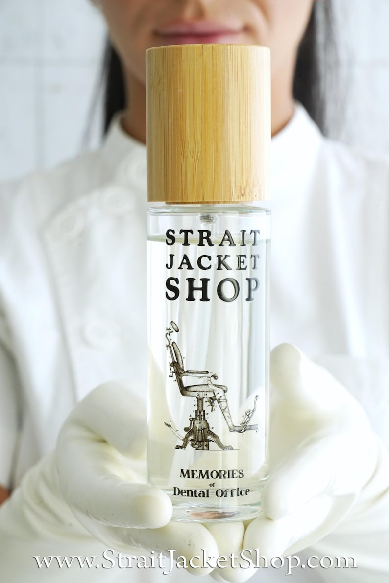 May include: A clear glass spray bottle with a light-coloured bamboo cap. The label features the text "STRAIT JACKET SHOP" above a black and white illustration of a dental chair, with the text "MEMORIES of Dental Office" printed below.