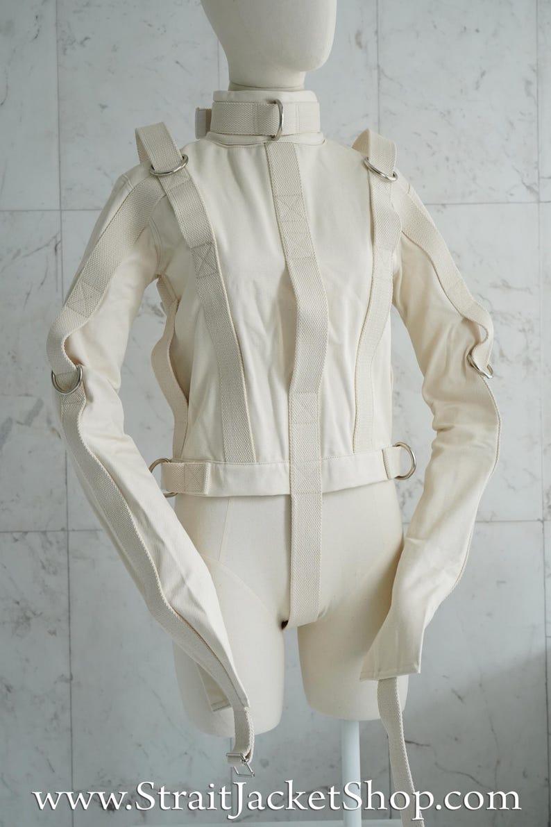 May include: A white canvas straitjacket with straps and buckles. The jacket has long sleeves that are tied at the wrists. The jacket is on a mannequin.