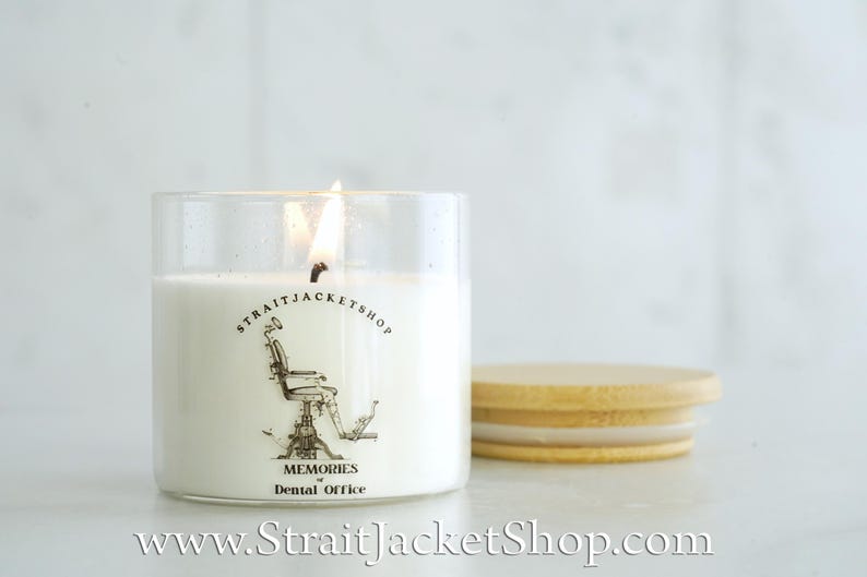 May include: A clear glass candle jar filled with white wax and a lit wick. The label features a black line drawing of a vintage dental chair, the text STRAITJACKETSHOP, and the words MEMORIES of Dental Office. A round bamboo lid sits nearby.