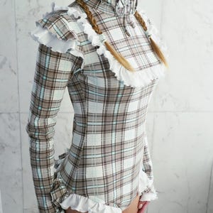 May include: A long-sleeved bodysuit in a plaid pattern of brown, white, and light blue. The garment features a high ruffled collar, ruffled shoulders, and ruffled trim at the wrists and legs. The website address www.StraitJacketShop.com is visible at the bottom.