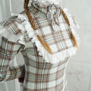 May include: A plaid dress with a high ruffled collar and long sleeves. The dress features a brown, white, and light blue plaid pattern. The website address www.StraitJacketShop.com is visible at the bottom of the image.