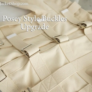 May include: Close-up of a cream-colored restraint garment with multiple straps and silver-colored buckles. The text "Posey Style Buckles Upgrade" is visible on the garment. The product appears to be made of a heavy-duty fabric.
