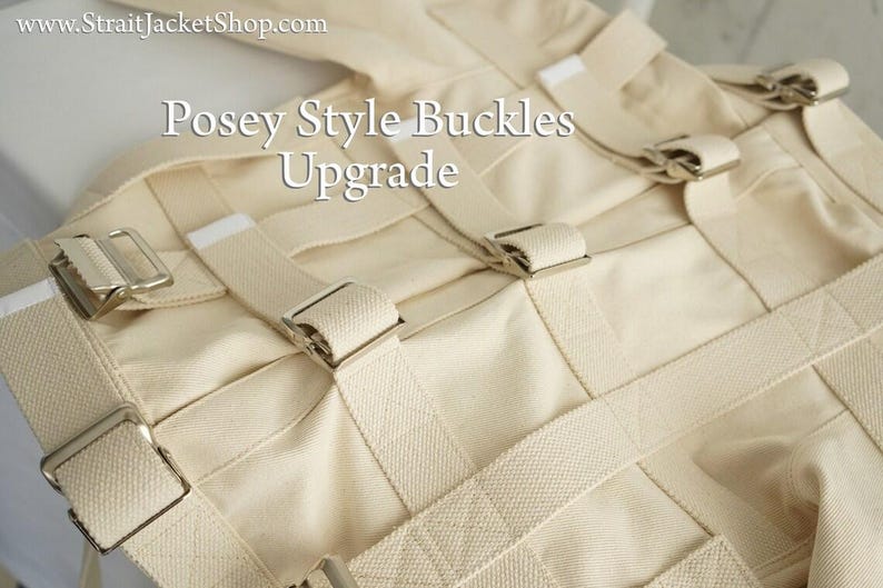 May include: Close-up of a cream-colored restraint garment with multiple straps and silver-colored buckles. The text "Posey Style Buckles Upgrade" is visible on the garment. The product appears to be made of a heavy-duty fabric.