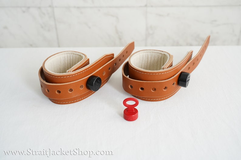 May include: Two brown leather wrist cuffs with white padded interiors and black metal buckles. A red metal key is also pictured.