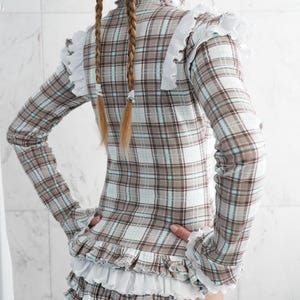 May include: A plaid long-sleeved top and shorts set in brown, white, and light blue. The top has a high collar and white ruffled trim at the shoulders and cuffs. The shorts feature multiple layers of white ruffles. The website address www.StraitJacketShop.com is visible.