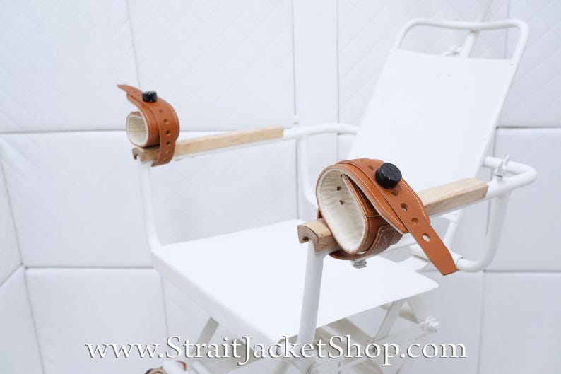 May include: White metal chair with brown leather restraints. The chair has a white padded seat and back. The restraints are attached to the arms of the chair.