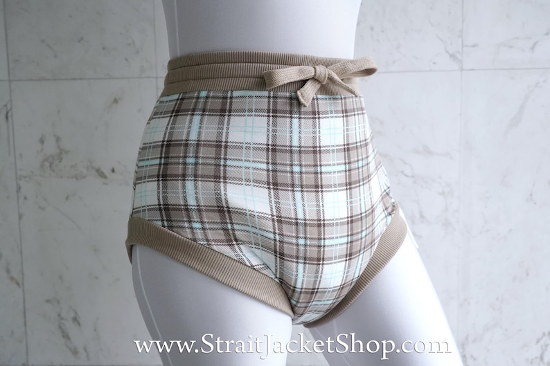 May include: High-waisted underwear with a plaid pattern in shades of brown, white, and light blue. The waistband and leg openings are trimmed with a solid tan fabric. A small bow detail is centered on the front of the waistband.