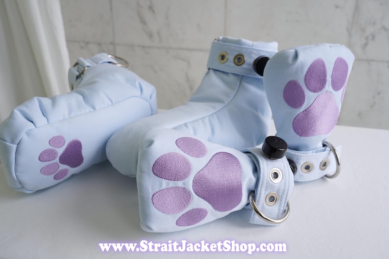 May include: A set of light blue fabric booties featuring embroidered purple paw prints on the front. Each bootie includes a cuff with silver-toned metal eyelets, a black circular fastener, and a metal D-ring. The text www.StraitJacketShop.com is visible.