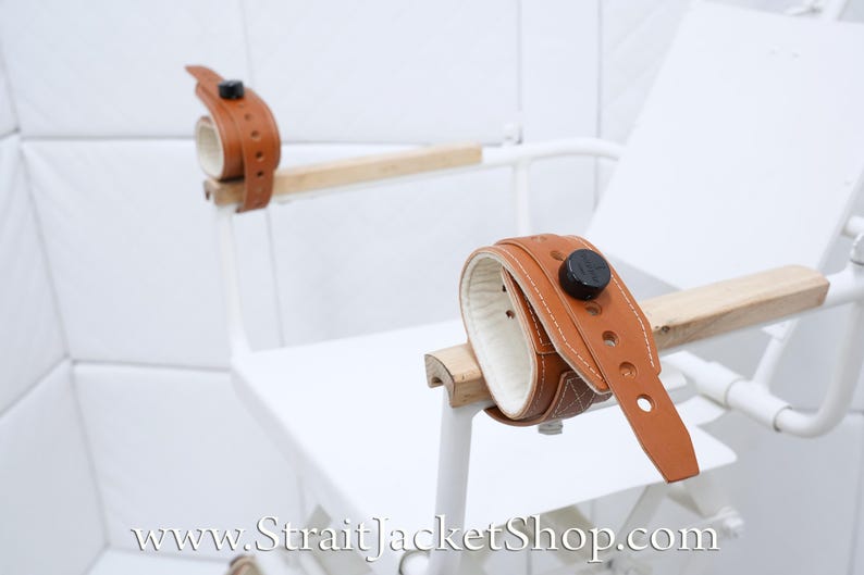 May include: Two brown leather restraints with metal buckles are attached to a white wooden chair. The chair is in a white room with white tile walls. The text "www.StraitjacketShop.com" is visible in the lower right corner of the image.