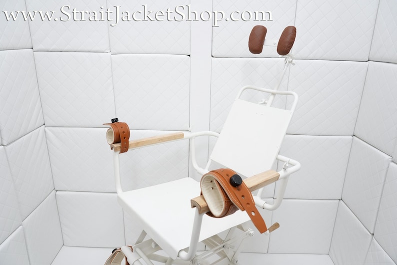 May include: White padded restraint chair with brown leather straps and headrests. The chair has a white padded seat and back, and the straps are adjustable. The chair is designed for restraint and is often used in medical settings.