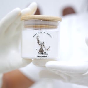 May include: A clear glass jar with a light-colored wooden lid containing a white substance. The label features a black and white illustration of a vintage dental chair with the text STRAITJACKETSHOP and MEMORIES of Dental Office.