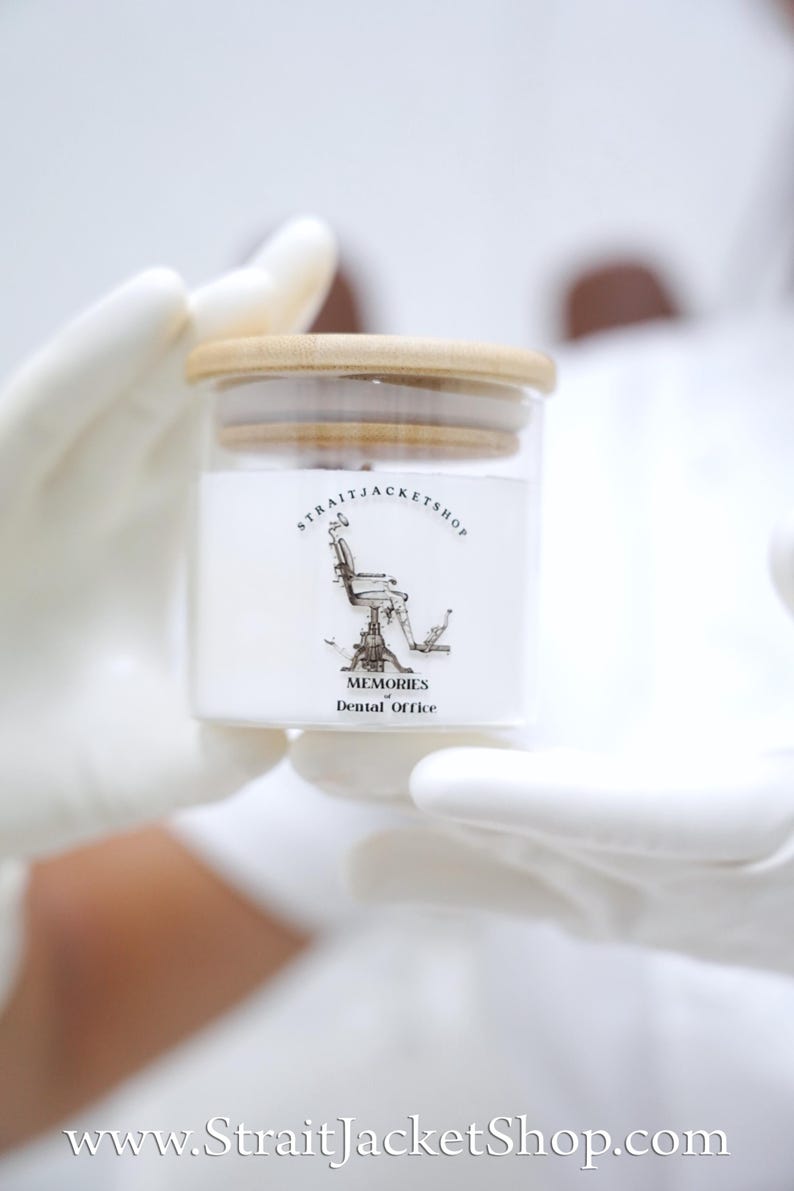 May include: A clear glass jar with a light-colored wooden lid containing a white substance. The label features a black and white illustration of a vintage dental chair with the text STRAITJACKETSHOP and MEMORIES of Dental Office.