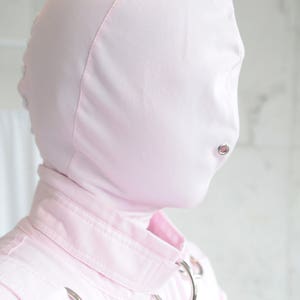 K&ouml;nnte beinhalten: Ein rosa Kapuzenkleidungsst&uuml;ck mit Rei&szlig;verschluss und zwei silbernen Metallringen an den Schultern. Die Kapuze hat eine kleine &Ouml;ffnung f&uuml;r den Mund.