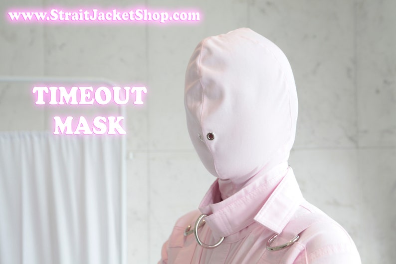 K&ouml;nnte beinhalten: Eine rosafarbene Stoffmaske mit einem einzigen Augenloch und einem silbernen Ring um den Hals. Die Maske ist mit "TIMEOUT MASK" beschriftet.