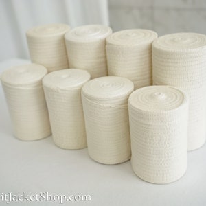 May include: A close-up of eight rolls of white elastic bandage, stacked in a pyramid shape on a white surface.
