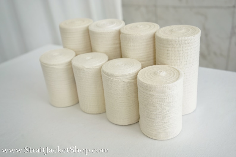 May include: A close-up of eight rolls of white elastic bandage, stacked in a pyramid shape on a white surface.