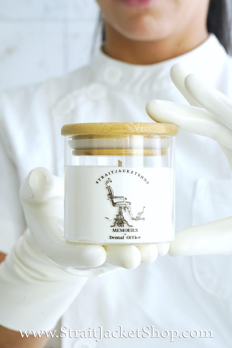 May include: A clear glass candle jar with a light-colored bamboo lid. The white label features a black line drawing of a vintage dental chair and the text STRAITJACKETSHOP MEMORIES of Dental Office in black ink on the front of the candle.