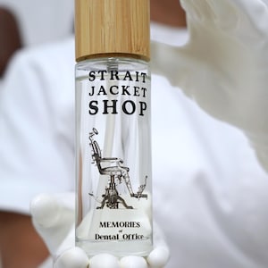May include: A clear glass spray bottle with a light brown bamboo cap. The bottle features black text reading STRAIT JACKET SHOP and MEMORIES of Dental Office, along with a detailed black line drawing of a vintage dental chair in the centre.