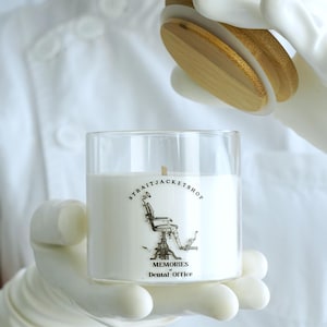 May include: A clear glass candle jar with a white wax fill and a wooden lid. The label features a black and white illustration of a dental chair with the text STRAITJACKETSHOP, MEMORIES of Dental Office, and the website StraitJacketShop.com.