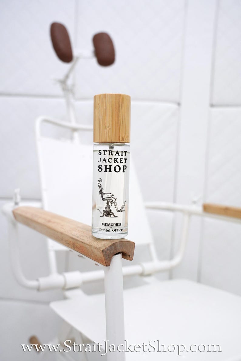 May include: A clear glass spray bottle with a light wood cap. The label features the text "STRAIT JACKET SHOP" above a black and white illustration of a dental chair, with the text "MEMORIES of Dental Office" printed below the illustration.