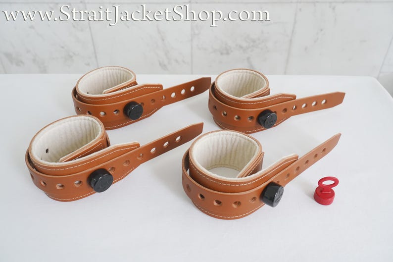 May include: Four brown leather wrist restraints with white padding and black buckles. The restraints are connected by a brown leather strap with holes for adjustment. A red plastic plug is also included.