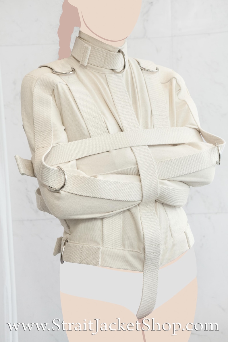 May include: A white canvas straitjacket with multiple straps and metal rings. The jacket is worn by a person with their arms crossed in front of them.