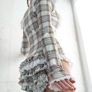 May include: A plaid top and skirt set in brown, white, and light blue. The long-sleeved top and skirt feature ruffled trim. The website address www.StraitJacketShop.com is visible at the bottom of the image.
