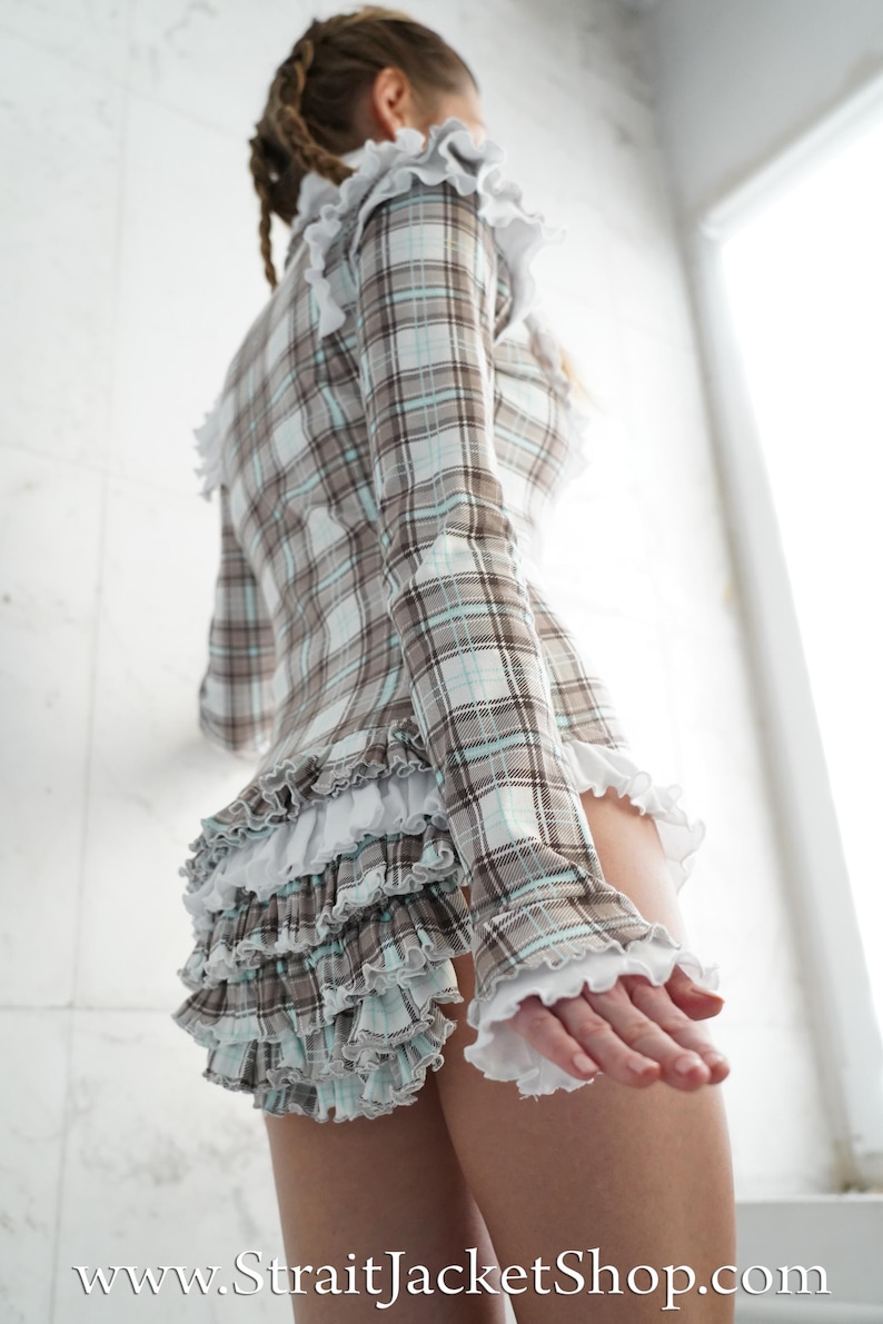 May include: A plaid top and skirt set in brown, white, and light blue. The long-sleeved top and skirt feature ruffled trim. The website address www.StraitJacketShop.com is visible at the bottom of the image.