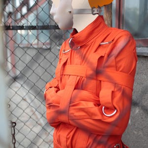 Op de afbeelding: Een oranje dwangbuis met meerdere riemen en metalen ringen. De jas wordt over zwarte kleding gedragen. Een wit gezichtsmasker en een hoofdsteun zijn ook zichtbaar. De website www.StraitJacketShop.com staat onderaan afgedrukt.