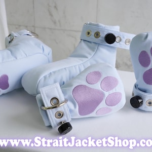 May include: Light blue padded footwear with purple paw print appliques, featuring adjustable straps with metal eyelets, silver D-rings, and black circular fasteners. The text www.StraitJacketShop.com is displayed at the bottom of the image.