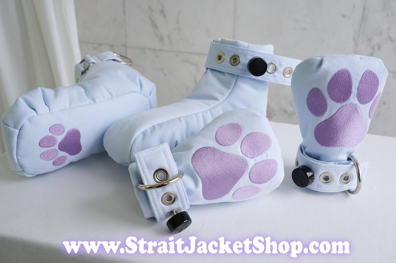May include: Light blue padded footwear with purple paw print appliques, featuring adjustable straps with metal eyelets, silver D-rings, and black circular fasteners. The text www.StraitJacketShop.com is displayed at the bottom of the image.